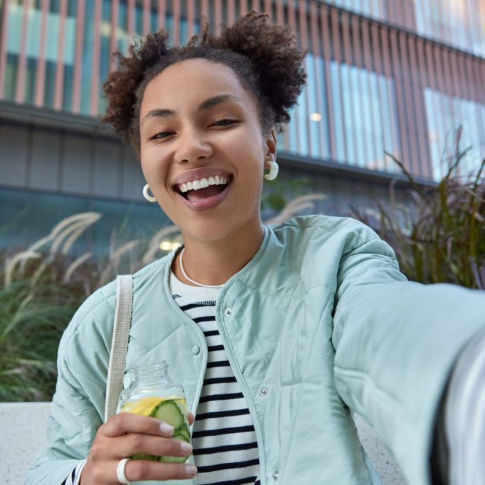 Happy optimistic girl with two hair buns dressed in jacket enjoys free time and walking in city holds bottle of detox drink makes selfie poses against modern building has fun during daytime.